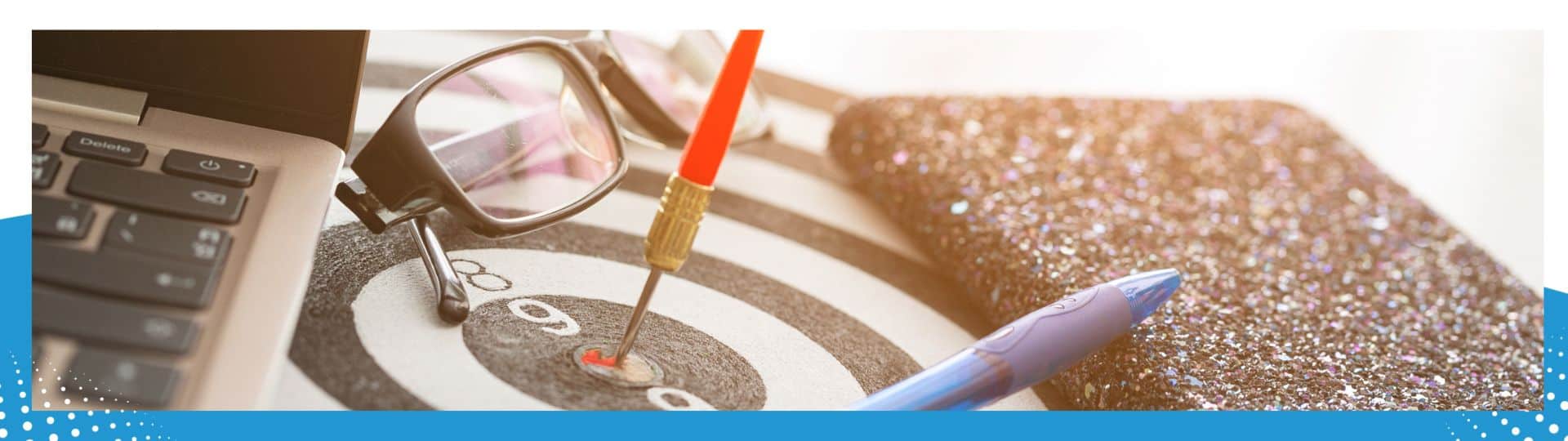 Close-up of a dart hitting the bullseye on a target board, surrounded by a laptop, glasses, and a pen, symbolizing precision, focus, and productivity in a professional setting.